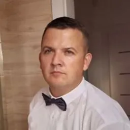 Portret mężczyzny w białej koszuli i muszce na tle ściany z beżowymi panelami.