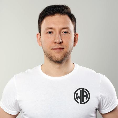 Portret mężczyzny w białej koszulce z okrągłym logo na piersi, na jednolitym szarym tle.