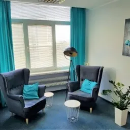 Jasny gabinet z dwoma ciemnymi fotelami typu uszak, małymi stolikami kawowymi, turkusowymi zasłonami i poduszkami, lampą podłogową oraz obrazem z motywem balonów na ścianie.