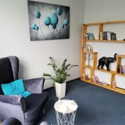 Jasny pokój z szarym fotelem uszakiem, drewnianym regałem z książkami i dekoracjami, obrazem z niebieskimi balonami na ścianie oraz rośliną w białej doniczce.