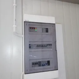 Tablica rozdzielcza z bezpiecznikami zamontowana na białej ścianie z widocznymi przewodami w białej rurce ochronnej.