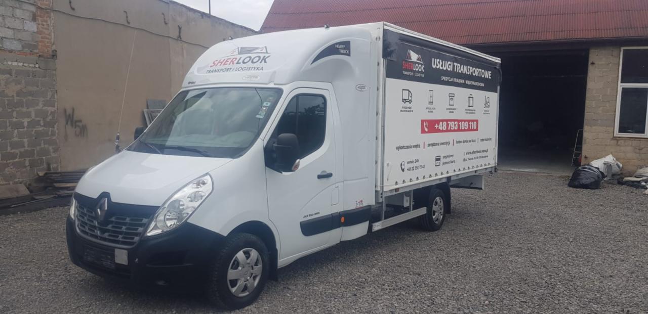 Biały bus Renault Master z zabudową plandekową, z logo firmy transportowej Sherlock, zaparkowany na żwirowym placu przed budynkiem gospodarczym.