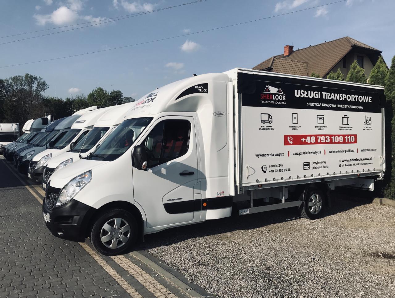 Flota białych samochodów dostawczych z reklamą usług transportowych i logistycznych, zaparkowana na żwirowym parkingu, w tle dom z brązowym dachem i błękitne niebo.