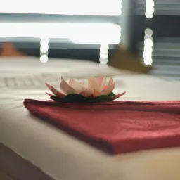 Przygotowane łóżko do masażu z czerwonym ręcznikiem i dekoracyjnym kwiatem lotosu w centrum, w tle rozproszone światło zza żaluzji.