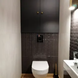 Nowoczesna toaleta podwieszana z szarą mozaiką na ścianie, szafką wiszącą z czarnymi frontami i złotymi gałkami oraz złotym oświetleniem ściennym.