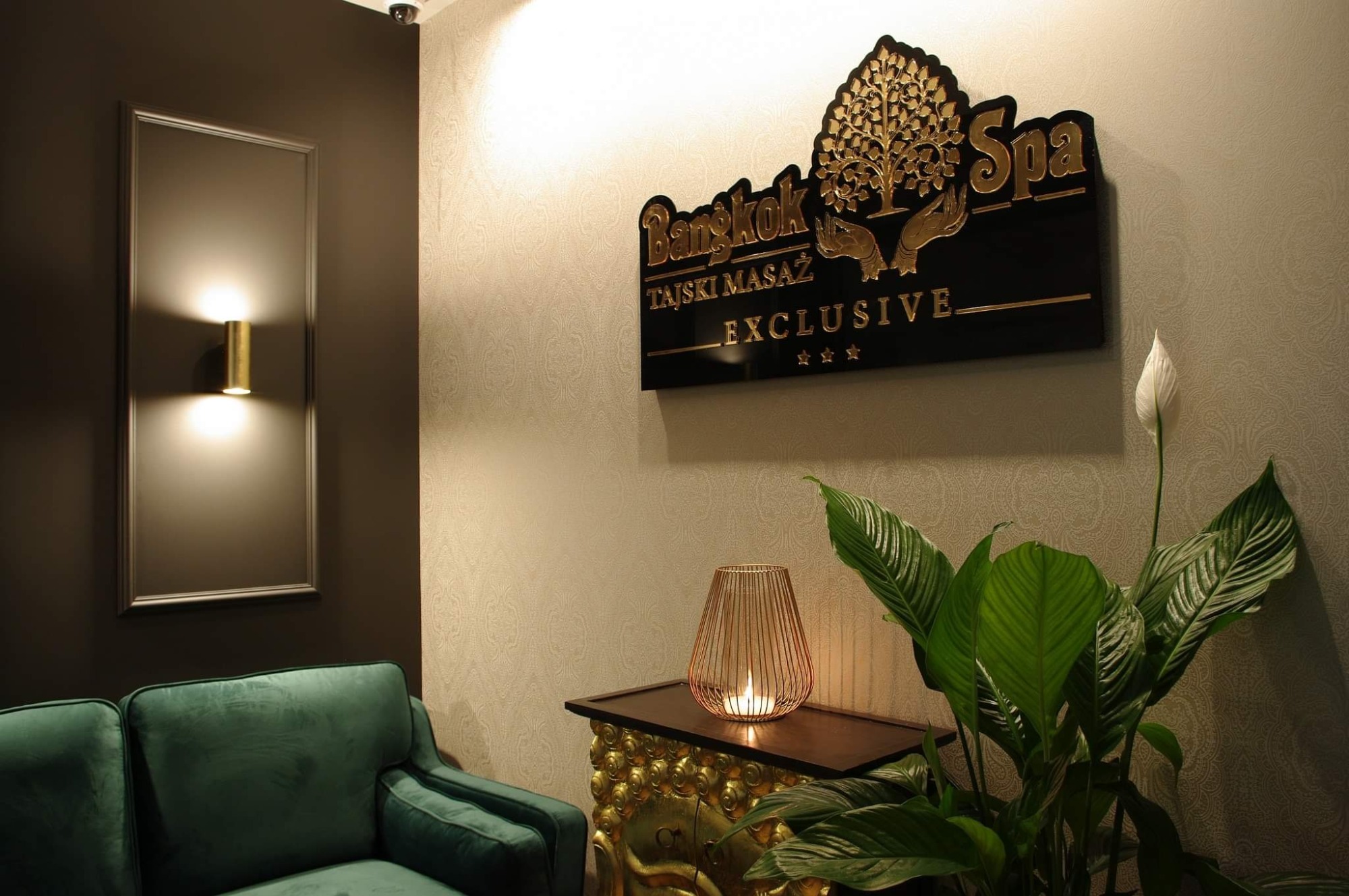 Recepcja salonu tajskiego masażu: zielona sofa, złoty kinkiet, stolik z dekoracyjną świecą i roślina doniczkowa na tle ściany z logo 'Bangkok Spa'.