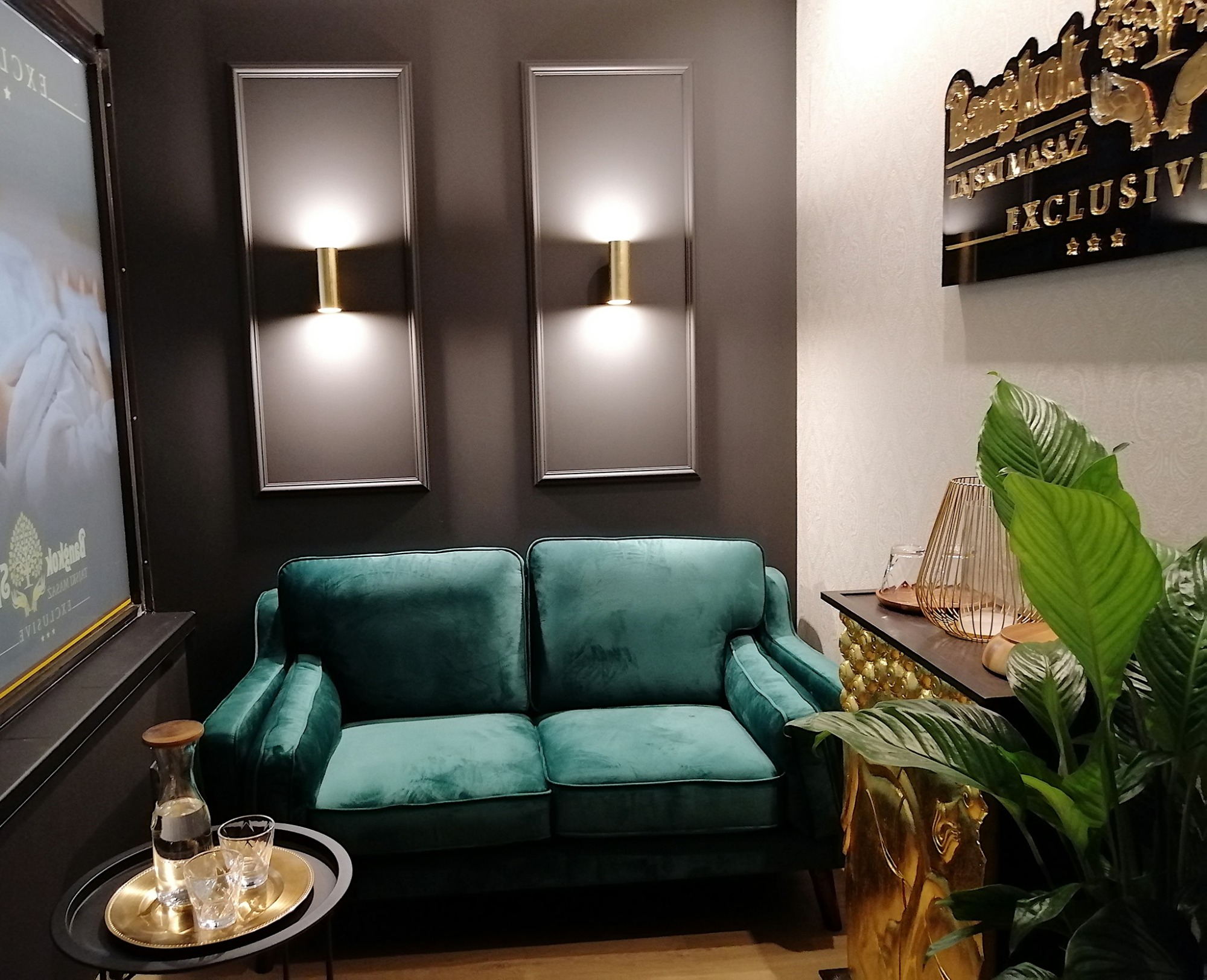 Szmaragdowa sofa z aksamitnym obiciem podświetlona przez dwa kinkiety w geometrycznych ramach; na stoliku obok karafka z wodą i szklanki na złotej tacy.