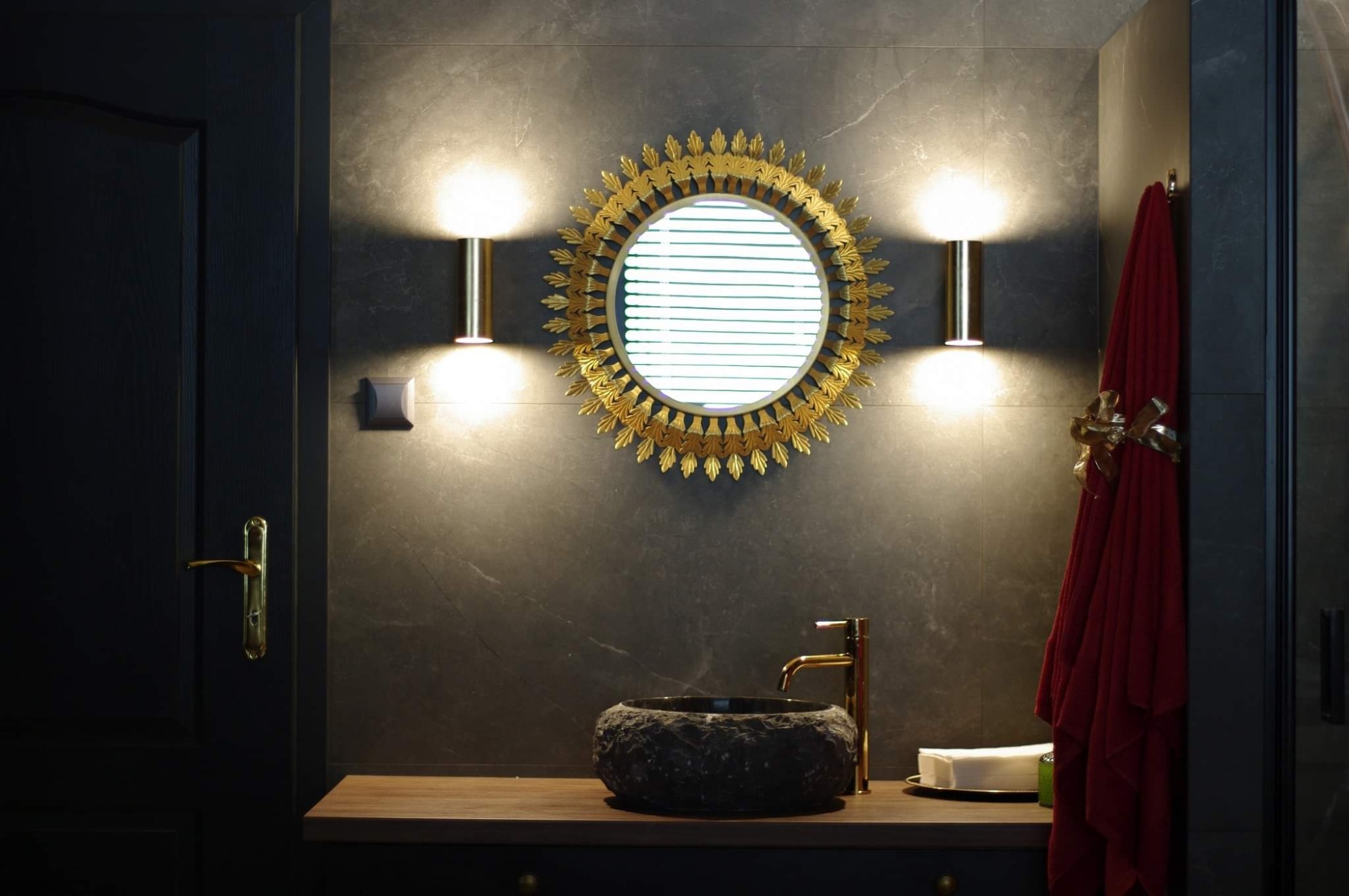 Nowoczesna łazienka z ciemnymi płytkami, okrągłym lustrem w złotej ramie, kamienną umywalką i złotą armaturą, oświetlona dwoma złotymi kinkietami.