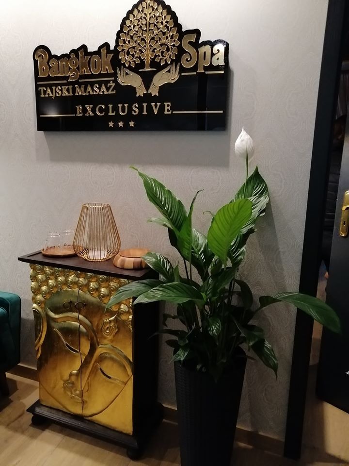 Recepcja salonu tajskiego masażu: złoty relief twarzy Buddy na szafce, roślina doniczkowa z białym kwiatem i czarny szyld z napisem 'Bangkok Spa Tajski Masaż Exclusive'.