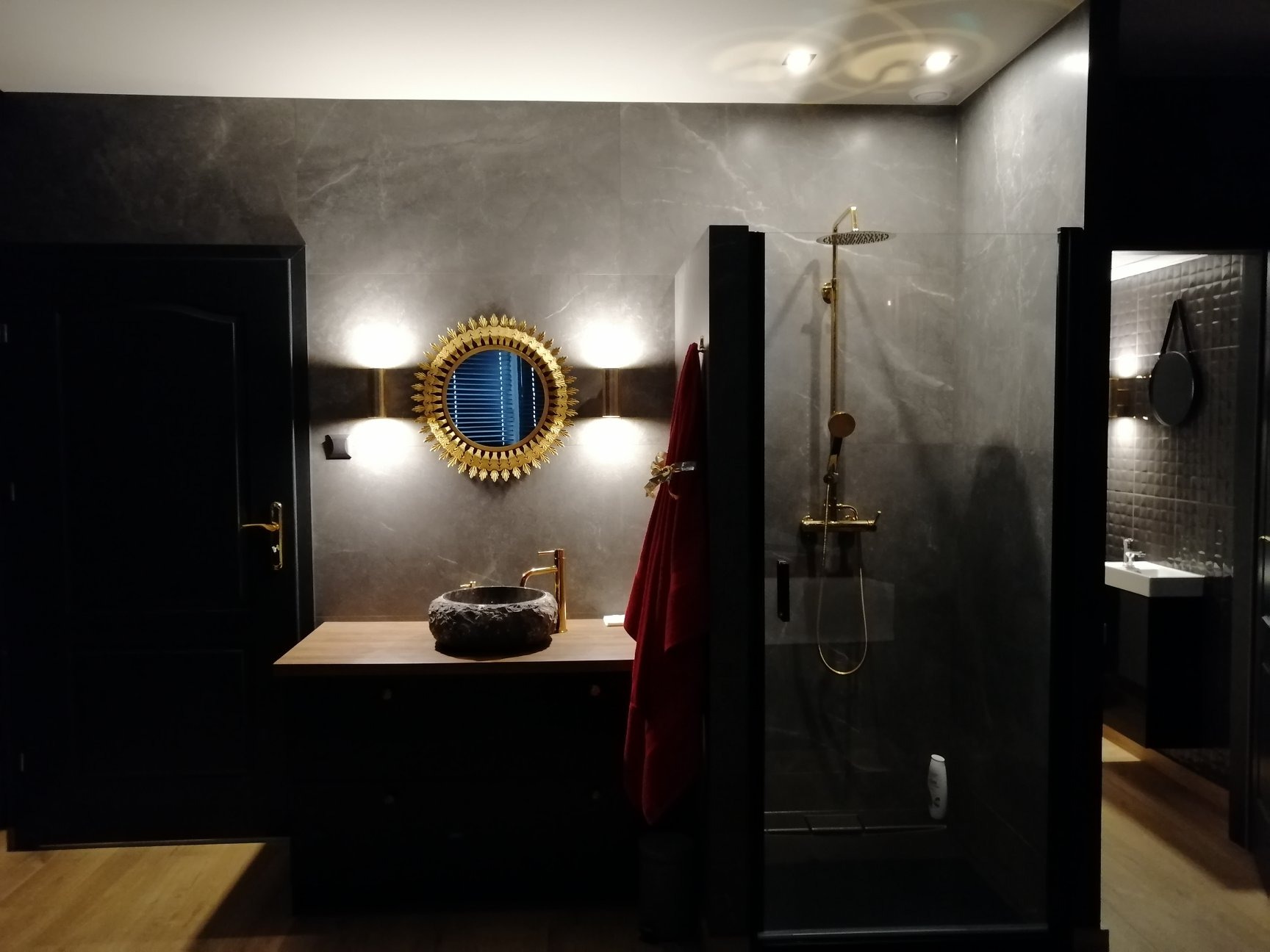 Ciemna łazienka z marmurowymi płytkami, okrągłym lustrem w złotej ramie, kamienną umywalką i złotą armaturą, kabina prysznicowa ze złotym zestawem natryskowym.