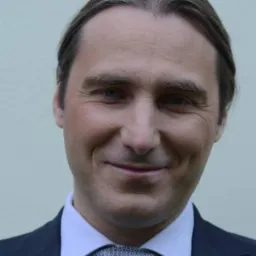 Portret uśmiechniętego mężczyzny w ciemnym garniturze, jasnej koszuli i wzorzystym krawacie na tle jednolitej ściany.