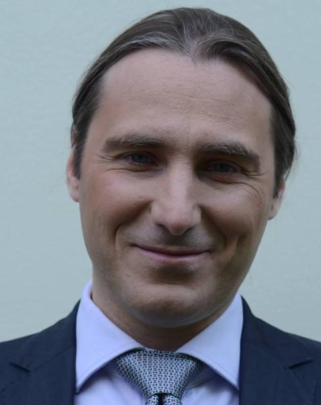 Portret uśmiechniętego mężczyzny w ciemnym garniturze, jasnej koszuli i wzorzystym krawacie na tle jednolitej ściany.