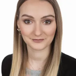 Portret uśmiechniętej kobiety z blond włosami, w czarnej marynarce, na białym tle.