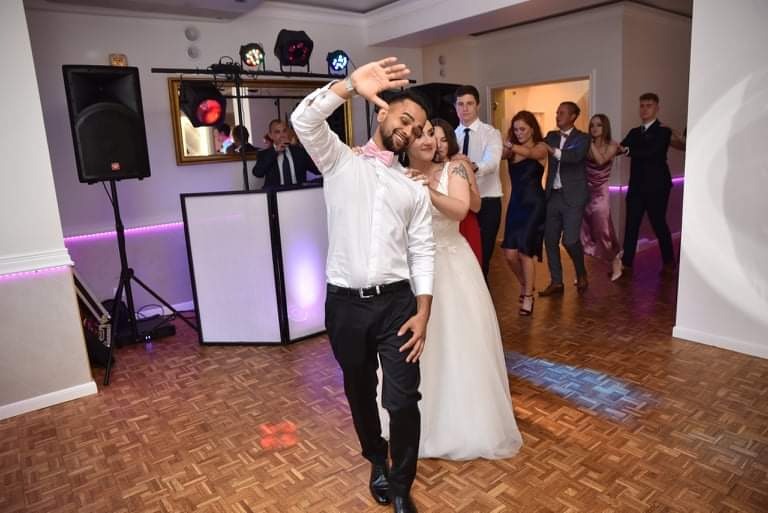 Para młoda prowadząca taneczny korowód gości weselnych, DJ grający w tle, oświetlenie dyskotekowe.