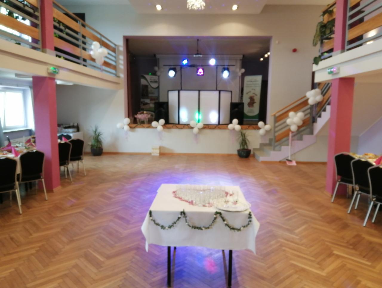 Sala bankietowa przygotowana na imprezę: scena z oświetleniem, dekoracje z białych balonów, stoły z nakryciami, parkiet z wzorem jodełki.