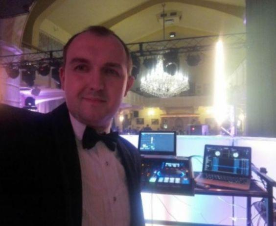 DJ w smokingu robiący selfie na tle sprzętu DJ-skiego i sali balowej z żyrandolem