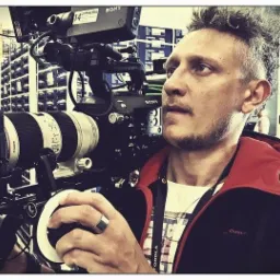 Operator kamery z profesjonalnym sprzętem Sony w magazynie, z widocznymi regałami w tle, ubrany w czarną kurtkę z czerwoną podszewką.