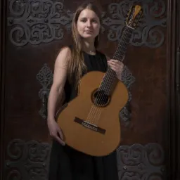 Młoda kobieta trzyma gitarę klasyczną na tle zdobionych, drewnianych drzwi.