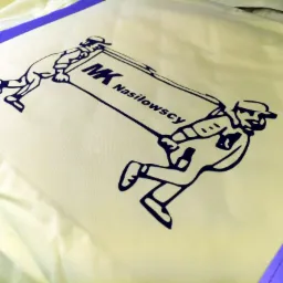 Żółta tkanina z nadrukowanym czarnym logo firmy MK Nasiłowscy, przedstawiającym dwóch mężczyzn w czapkach niosących szyld z nazwą firmy.