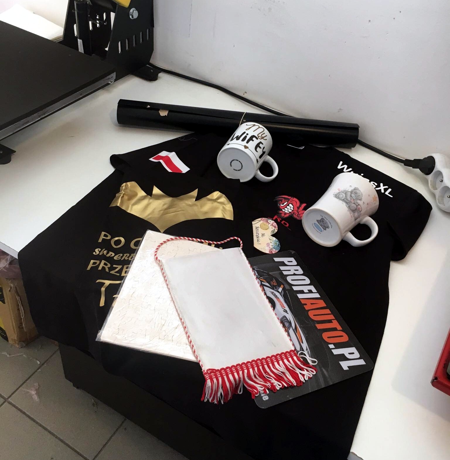 Czarne koszulki z nadrukami (złoty symbol nietoperza, napis) oraz kubki z nadrukami, leżące na stole obok prasy termotransferowej i innych akcesoriów.