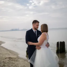 Para młoda obejmująca się na plaży, w tle woda i pochmurne niebo, widoczne drewniane pale w wodzie, ona w białej sukni z koronkowymi detalami, on w granatowym garniturze.