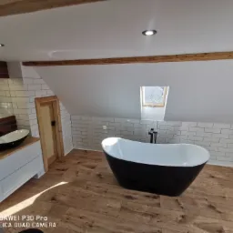 Nowoczesna łazienka na poddaszu z wolnostojącą czarno-białą wanną, białą umywalką w kształcie misy na drewnianym blacie i białymi kaflami na ścianach.