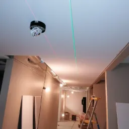 Montaż sufitu podwieszanego: widoczny laser krzyżowy wyznaczający poziom, płyty na podłodze, drabina i oświetlenie budowlane w tle.
