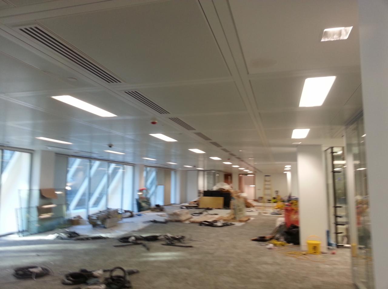 Rozległe biuro w trakcie prac wykończeniowych; widoczne rozłożone narzędzia, materiały budowlane, odsłonięta podłoga i sufit z panelami oraz oświetleniem.