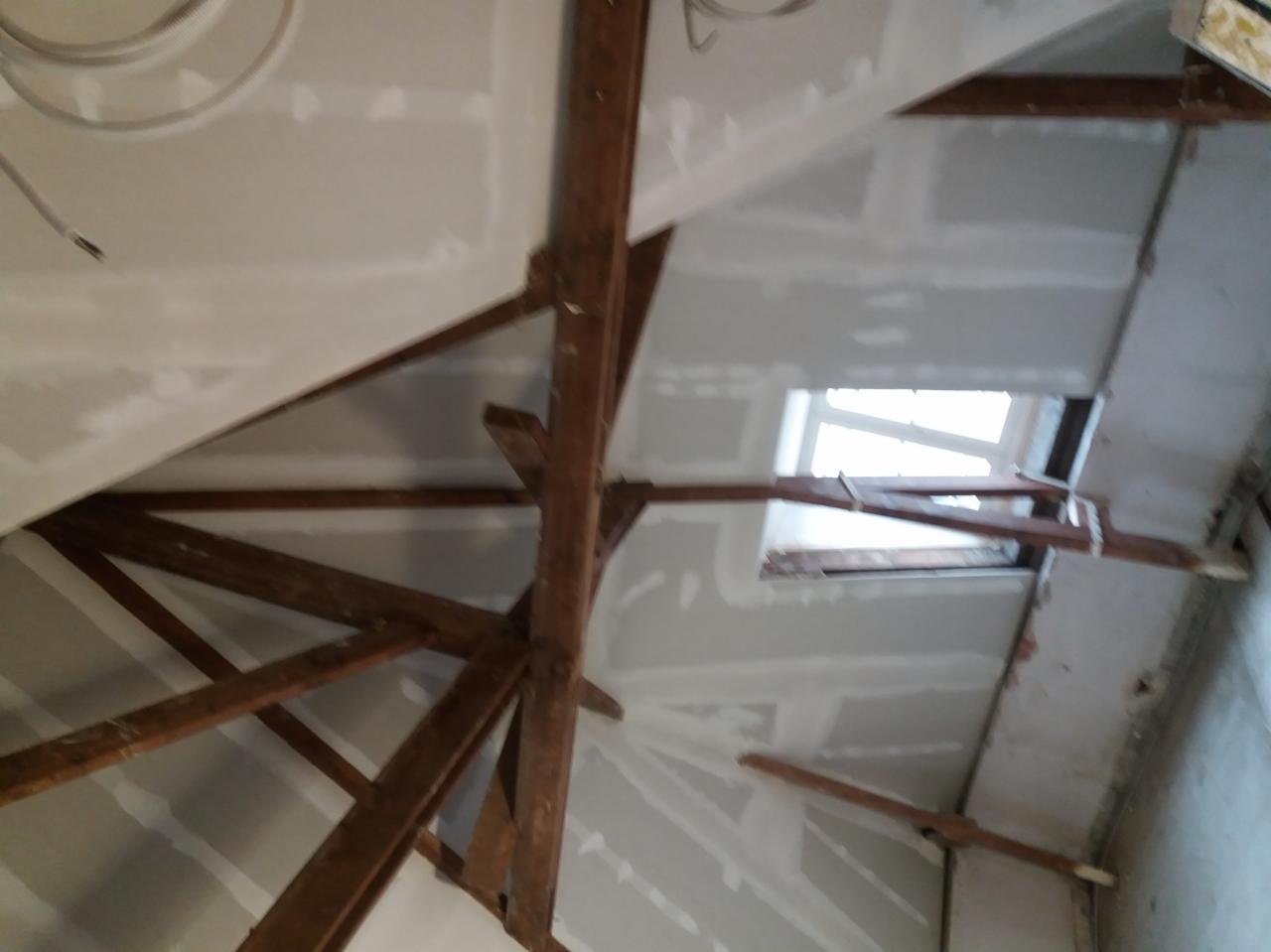 Poddasze z widoczną konstrukcją drewnianą i zamontowanymi płytami na ścianach i suficie, częściowo zaszpachlowane, widoczne okno dachowe.