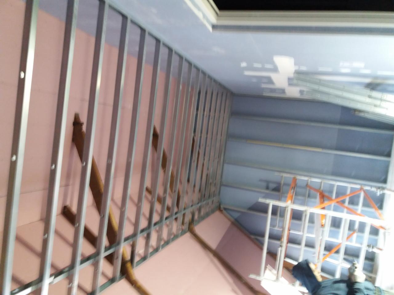 Montaż konstrukcji metalowej na różowej ścianie, częściowo pokrytej płytami, widoczny fragment sufitu i drabina w tle.