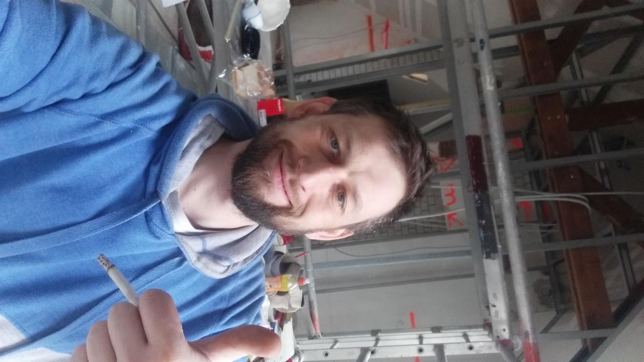 Mężczyzna z papierosem na tle konstrukcji sufitu podwieszanego z metalowych profili, widoczne przewody elektryczne i drabina.