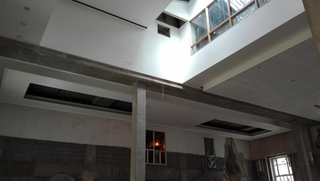 Wnętrze w trakcie prac wykończeniowych: widoczne otwory w suficie, surowy beton, ściany przygotowane do malowania, okna dachowe z drewnianą ramą i częściowo zakryte folią okna.