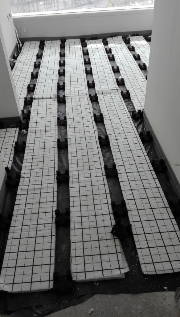 Przygotowanie podłoża pod ogrzewanie podłogowe: białe maty grzewcze z czarną siatką na czarnej folii izolacyjnej, widoczne czarne elementy mocujące.