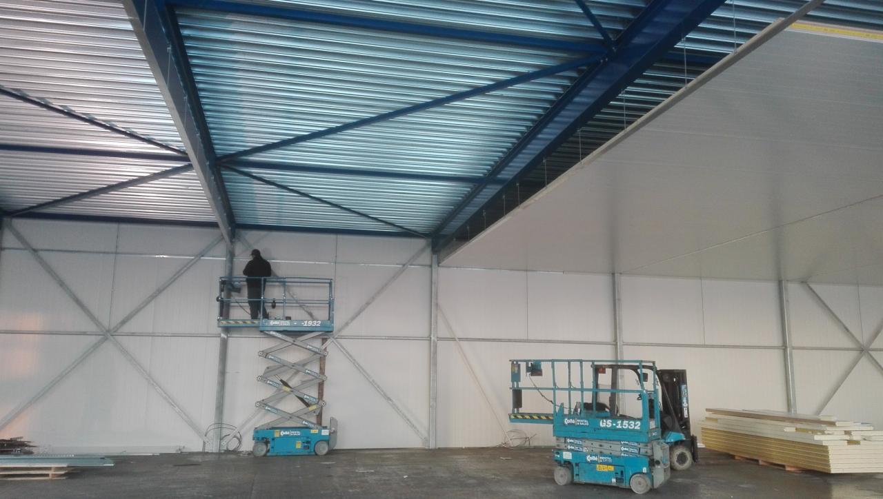 Montaż płyt na dużej powierzchni sufitu w hali magazynowej, widoczny pracownik na podnośniku koszowym oraz elementy konstrukcji dachu i ścian.