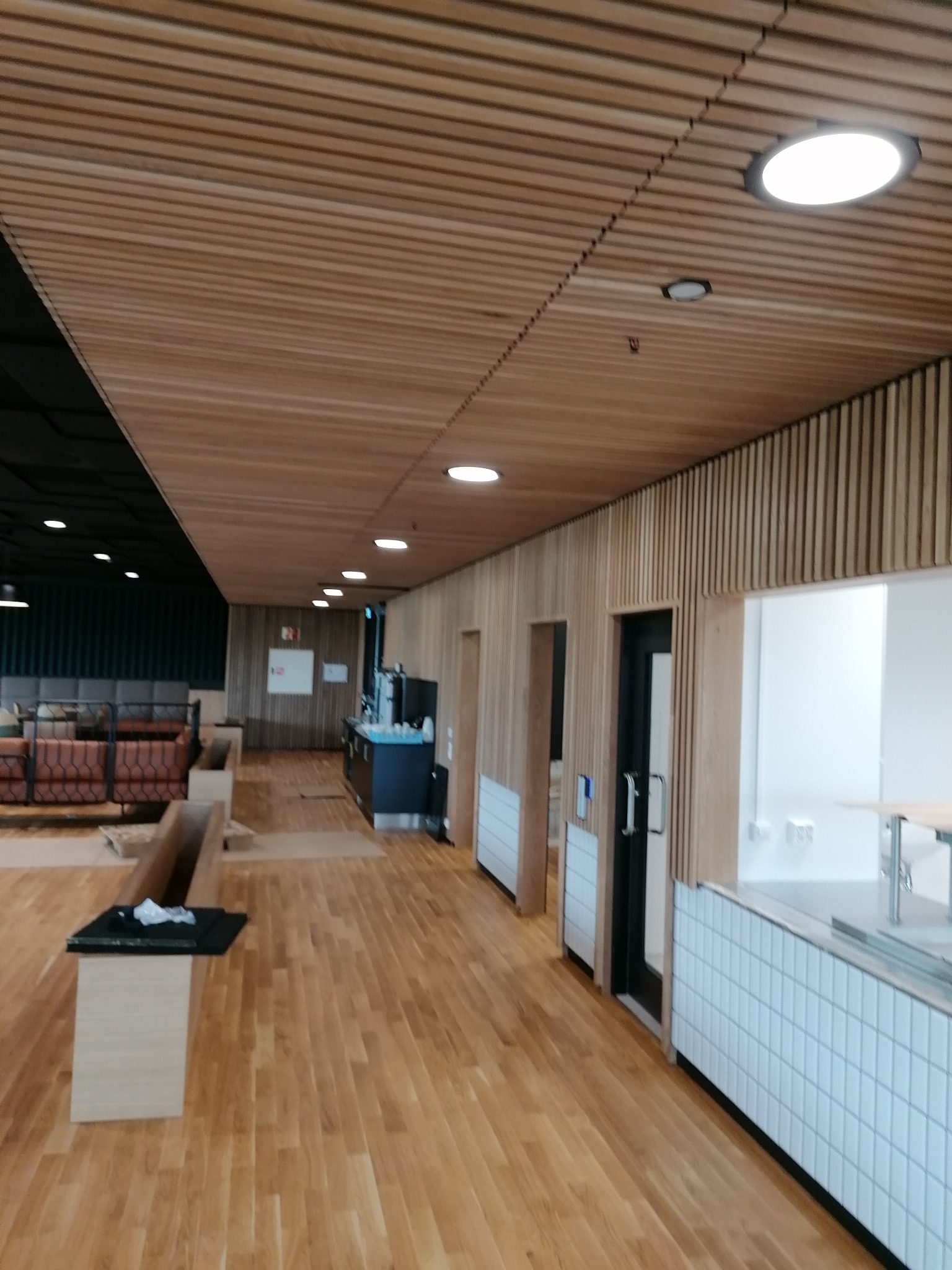 Wnętrze komercyjne z drewnianą podłogą, sufitem z drewnianych listew i ścianami wykończonymi panelami. Widoczne oświetlenie sufitowe, meble oraz fragment wyposażenia kuchni. Część sufitu wykończona...