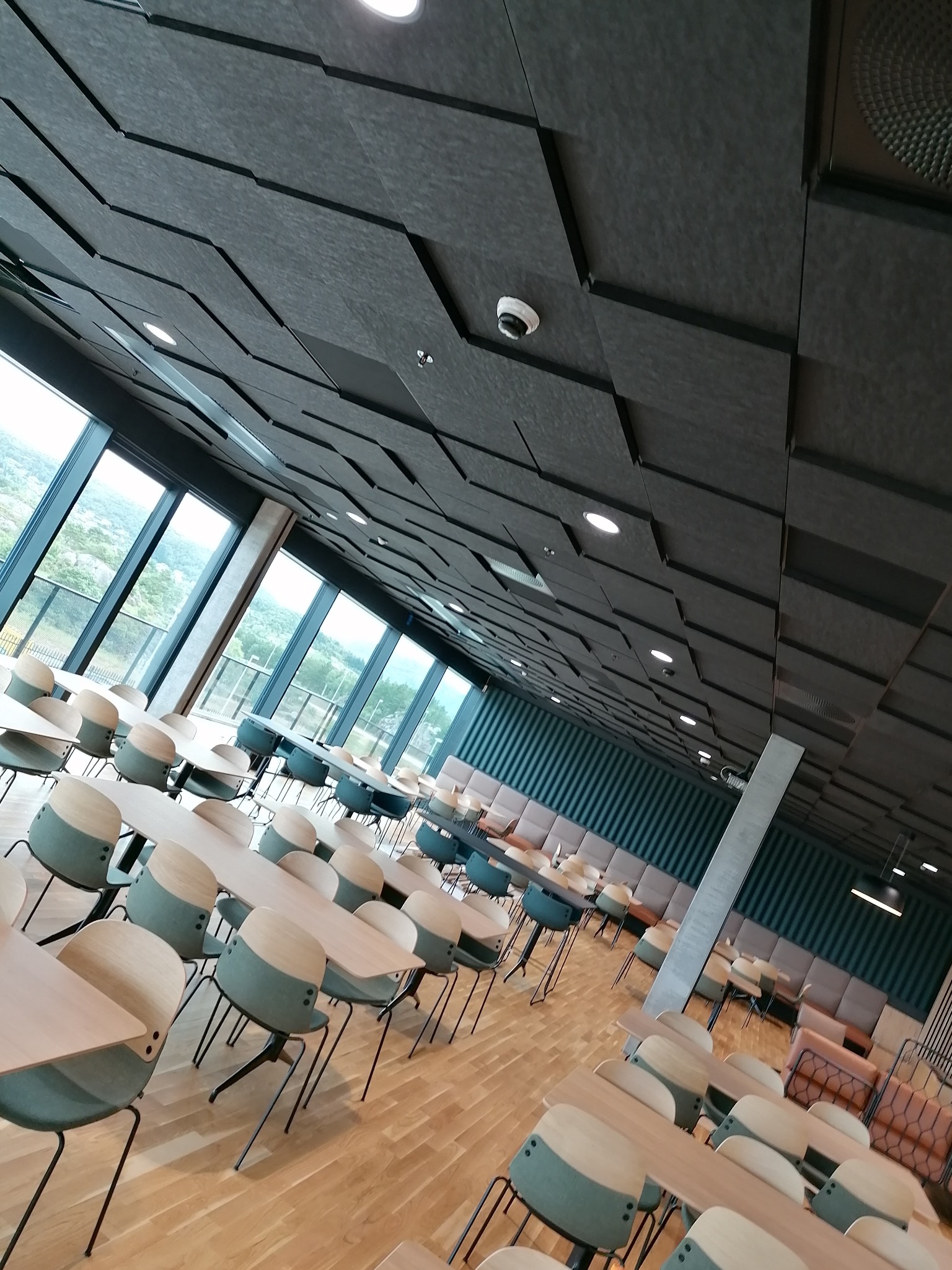 Duża sala konferencyjna z geometrycznym sufitem z paneli akustycznych, widok na zewnątrz przez panoramiczne okna, stoły i krzesła ustawione w rzędach, oświetlenie punktowe i dekoracyjne.