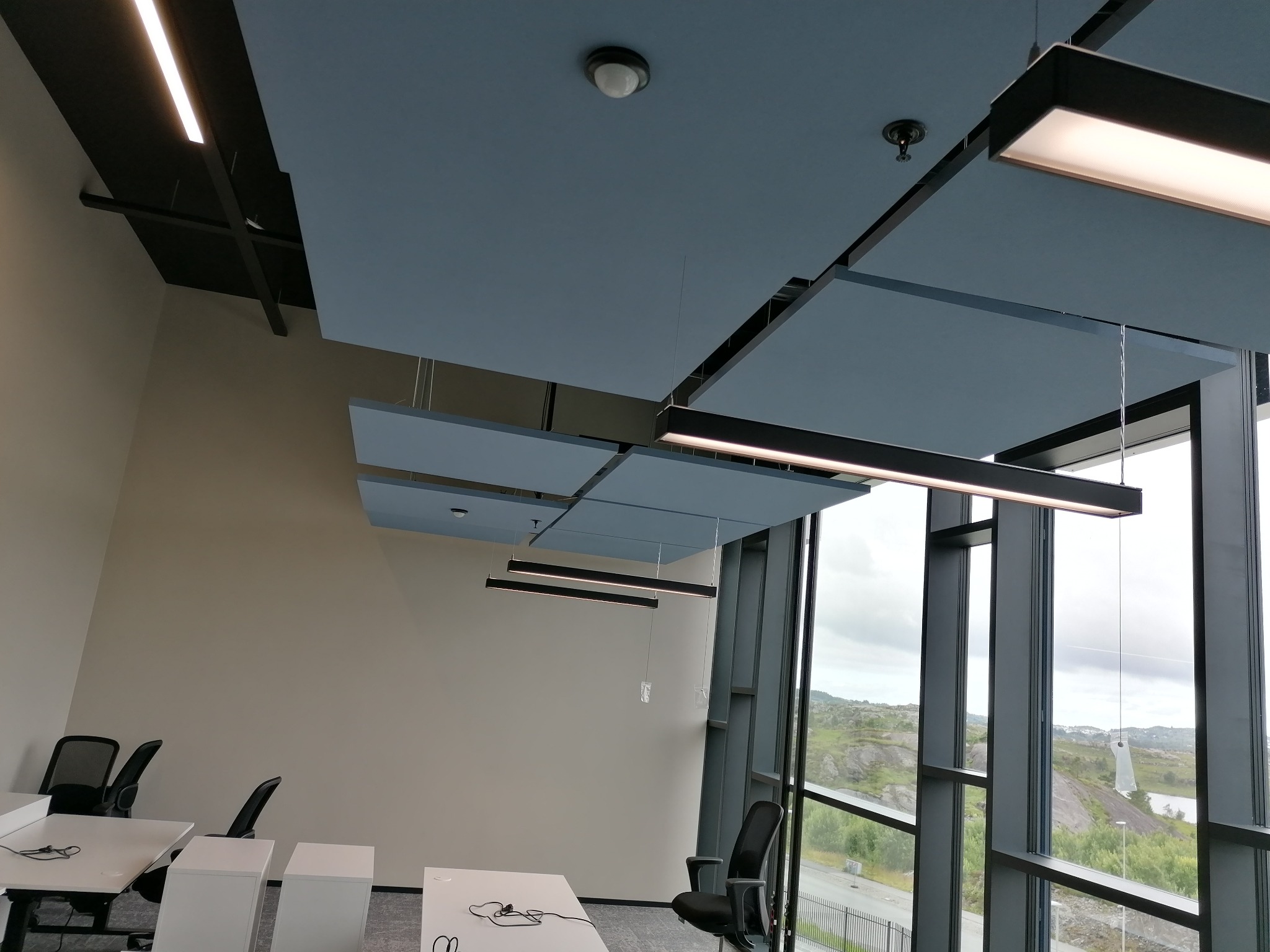 Biuro z akustycznymi panelami sufitowymi w kolorze gołębiej szarości, podwieszanymi lampami liniowymi i widokiem na wzgórza za oknem.