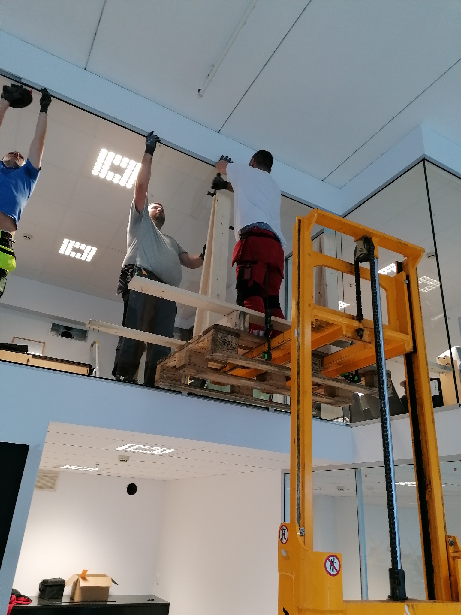 Trzech mężczyzn w strojach roboczych montuje sufit napinany w biurze, używając podnośnika paletowego i platformy roboczej, widoczne oświetlenie sufitowe i fragmenty szklanych ścianek działowych.