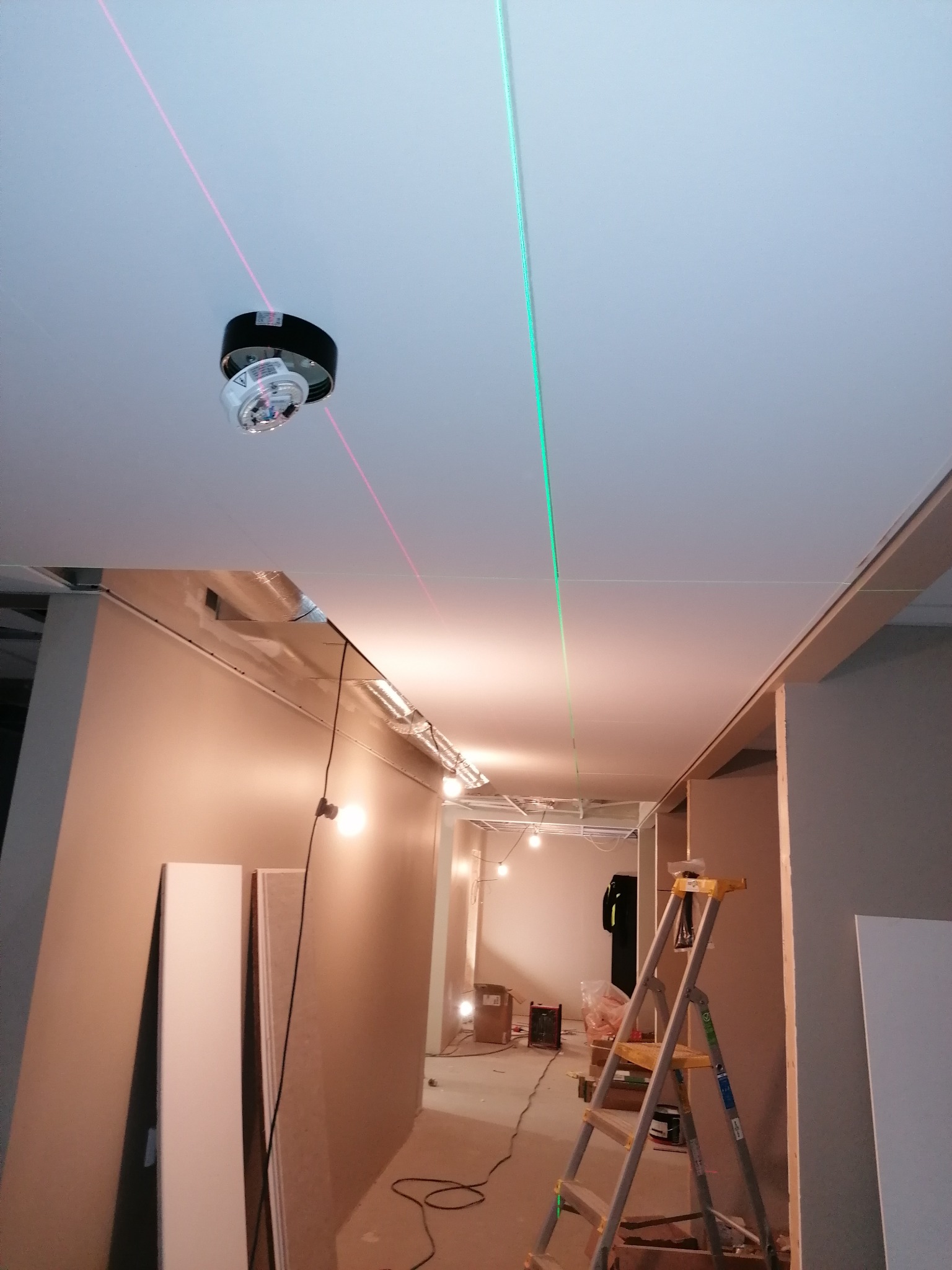 Montaż sufitu podwieszanego: widoczny laser krzyżowy wyznaczający poziom, płyty na podłodze, drabina i oświetlenie budowlane w tle.