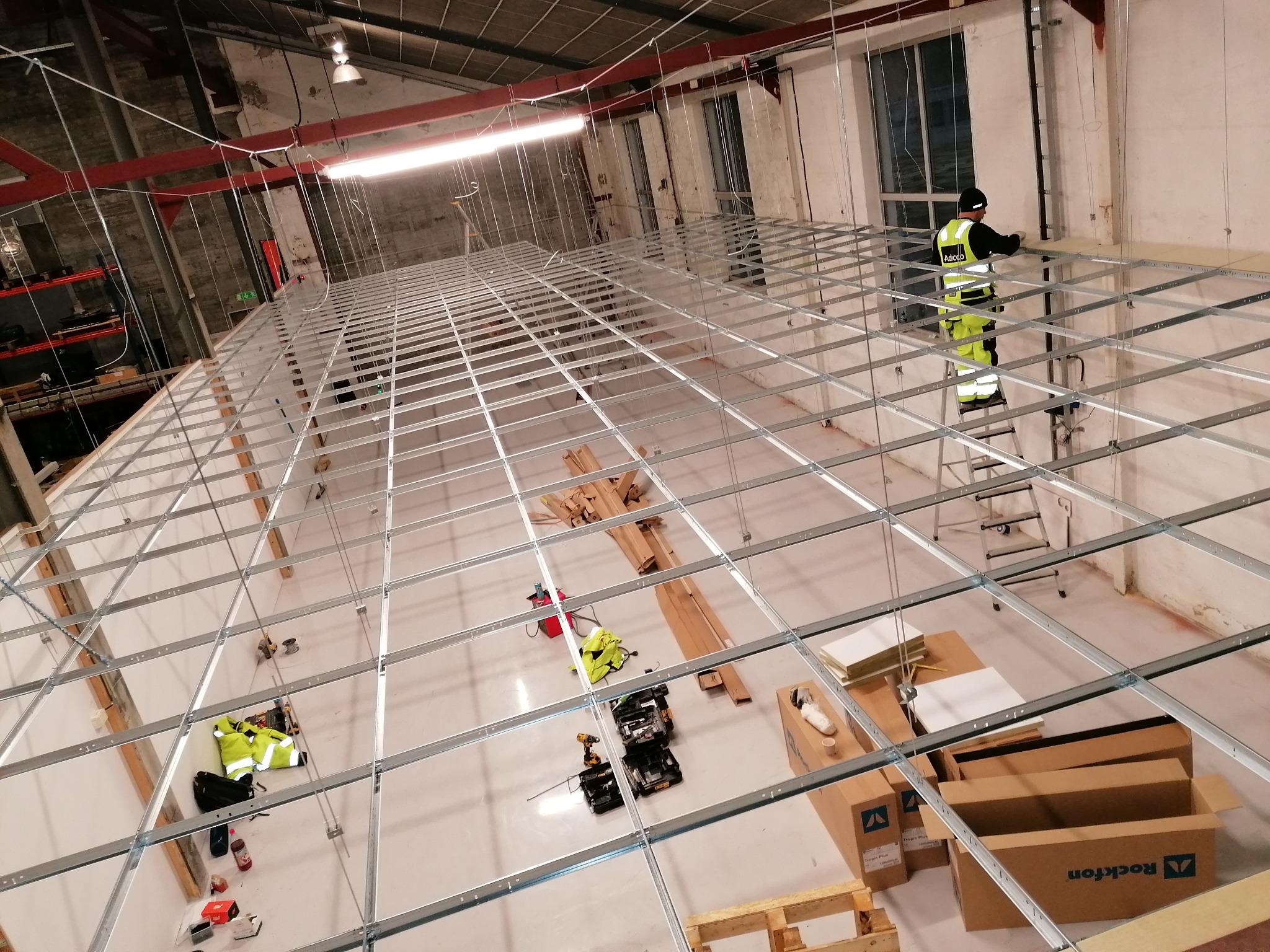 Montaż metalowego rusztu sufitowego w Sanoku, widok z góry na pracownika na drabinie i przygotowane materiały budowlane.