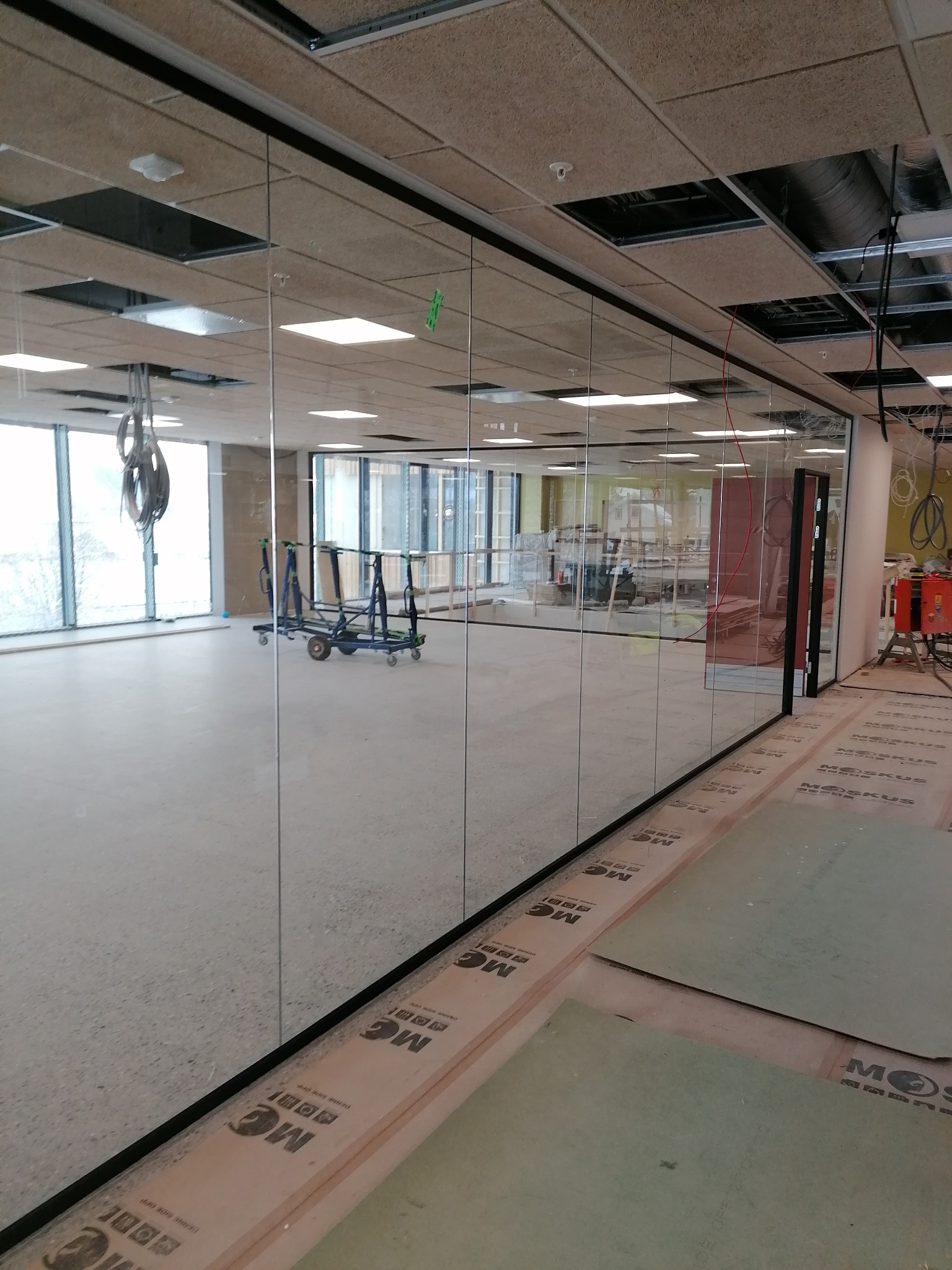 Montaż szklanych ścian działowych w biurze, widoczne profile, sufit z paneli akustycznych, w tle wózek transportowy i narzędzia.