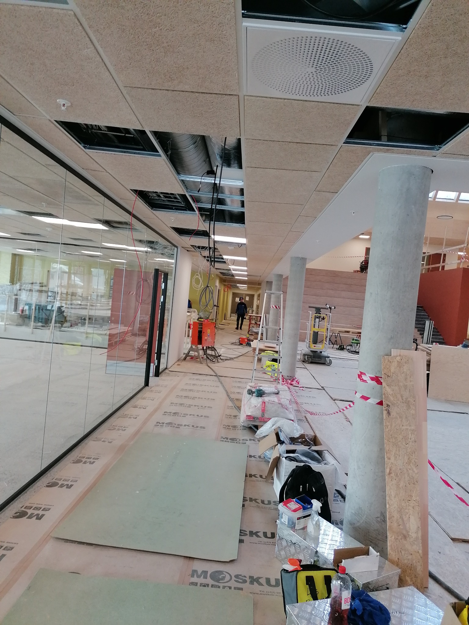 Montaż sufitu podwieszanego z płyt o strukturze drewna w dużym pomieszczeniu biurowym, widoczne elementy konstrukcyjne, otwory na instalacje, narzędzia i materiały budowlane na podłodze...