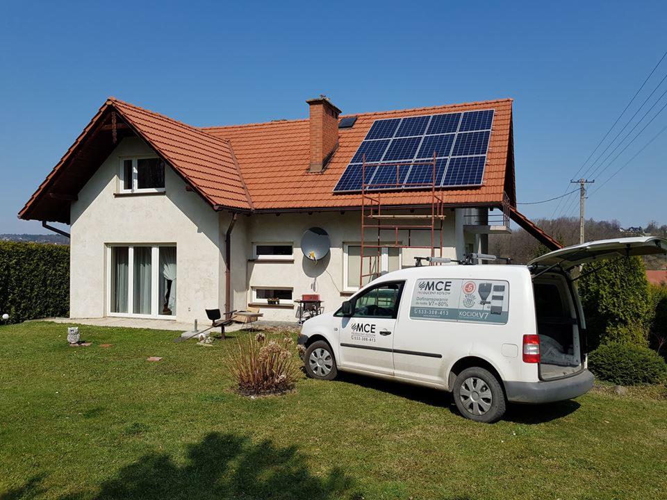 Dom jednorodzinny z panelami słonecznymi na dachu, drabina oparta o dach, biały samochód dostawczy z otwartym bagażnikiem i logo firmy na boku, słoneczny dzień.