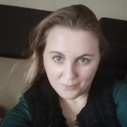 Portret kobiety z jasnymi włosami i niebieskimi oczami, ubrana w zieloną bluzkę i czarną kamizelkę z futerka, na rozmytym tle.