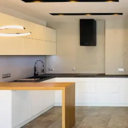 Nowoczesna kuchnia z białymi szafkami, drewnianym blatem wyspowym i czarnym okapem, oświetlona listwami LED.