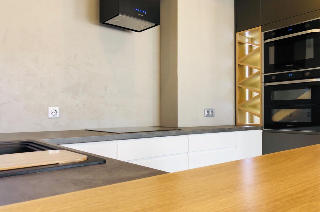 Nowoczesna kuchnia: białe szafki, szary blat, drewniana wyspa, czarny okap i wbudowane piekarniki Samsung obok podświetlanego regału na wino.
