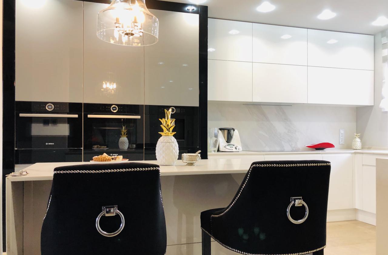Nowoczesna kuchnia z błyszczącymi frontami, wbudowanymi piekarnikami Bosch, wyspą z blatem z konglomeratu i dwoma czarnymi krzesłami barowymi z ozdobnymi srebrnymi nitami.
