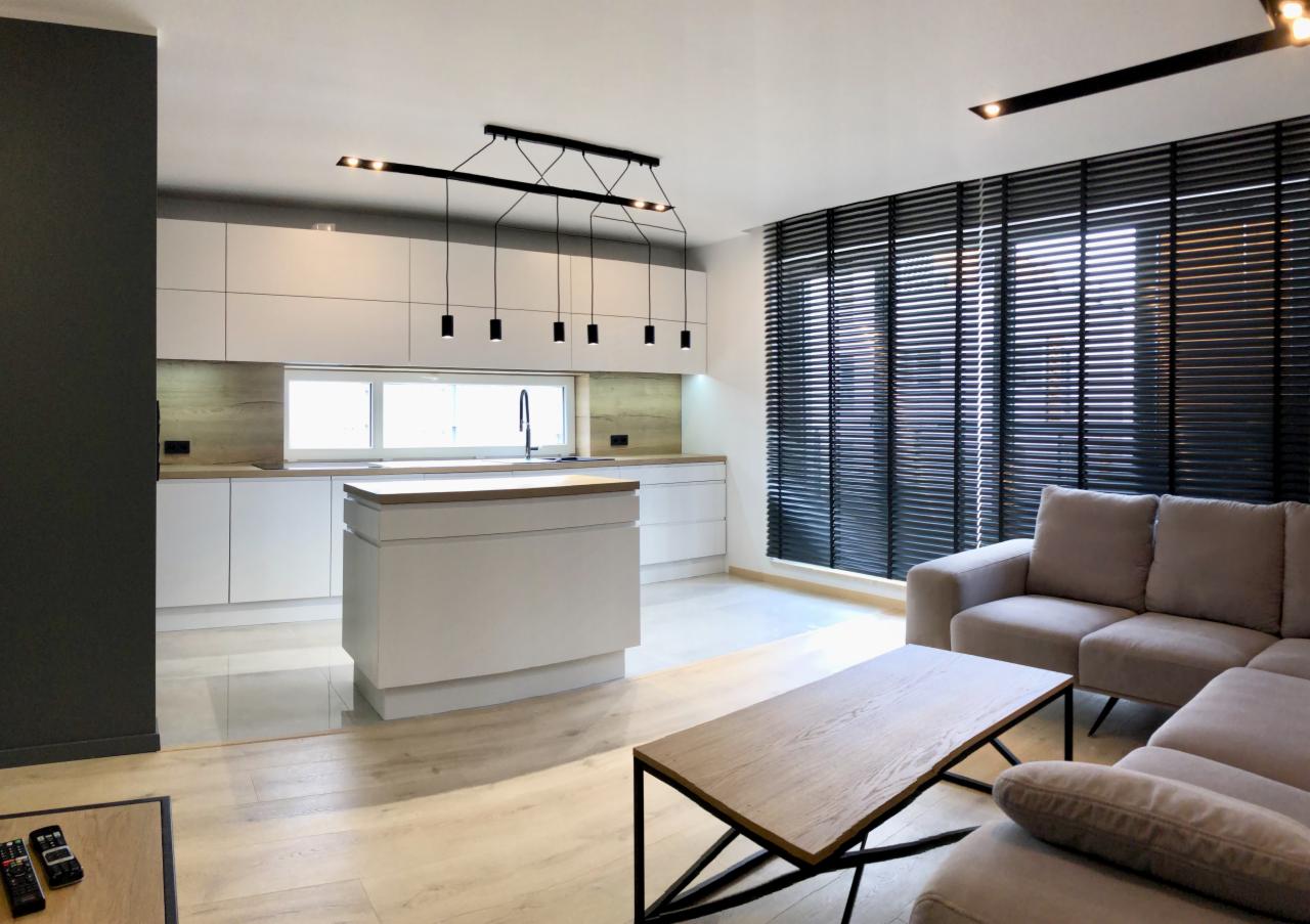 Nowoczesna, biała zabudowa kuchenna z wyspą, oświetlona wiszącymi lampami, widok z salonu z kanapą i stolikiem kawowym, żaluzje w oknach.