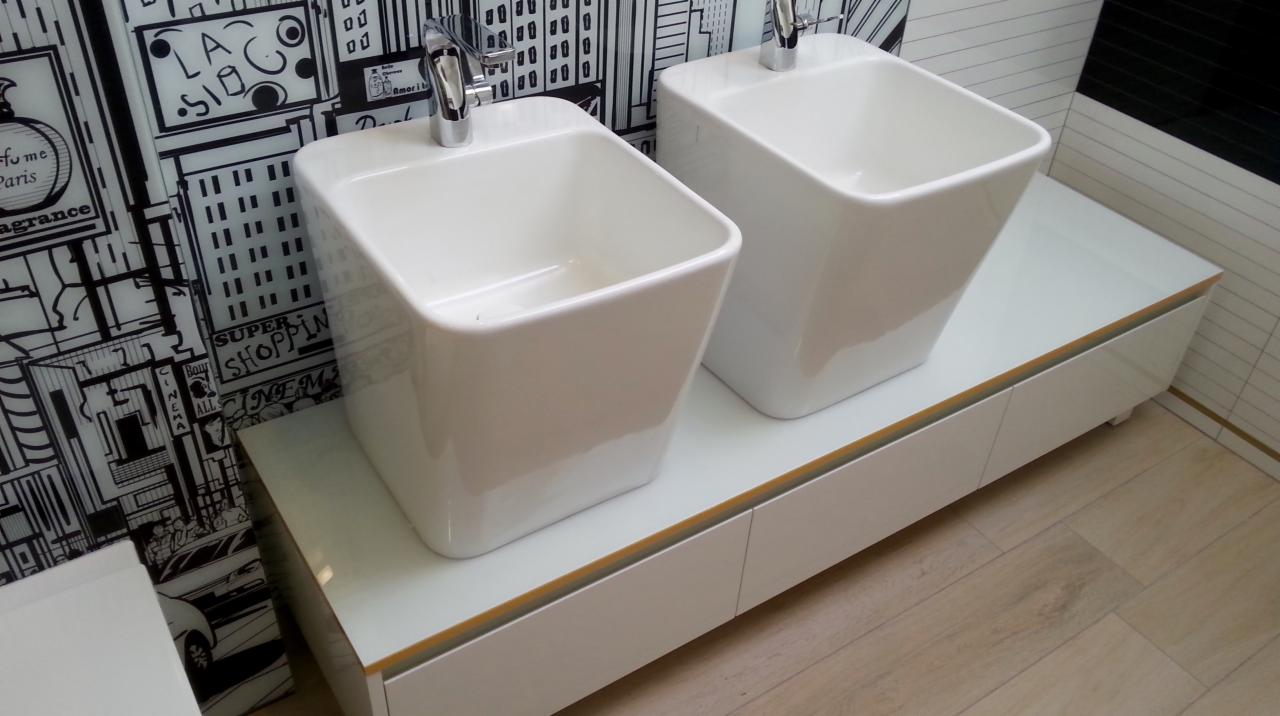 Dwie białe, kwadratowe umywalki nablatowe z chromowanymi bateriami na białej szafce z subtelną złotą lamówką, na tle ściany z czarno-białym motywem miejskim.