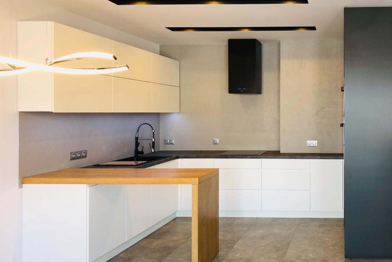 Nowoczesna kuchnia z białymi szafkami, drewnianym blatem wyspowym i czarnym okapem, oświetlona listwami LED.